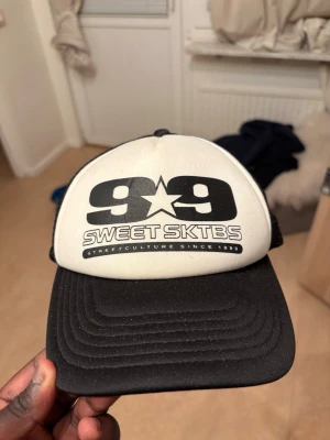 Sweet SKTBS svartvit truckerkeps - Säljer en svartvit truckerkeps från Sweet SKTBS med stort 99-tryck och stjärna framtill. Kepsen har mesh på sidorna och bak, böjd skärm och justerbar snapback. Perfekt för dig som gillar streetstyle och skatekultur.
