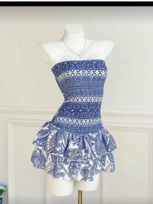 Blåvit axelbandslös kortklänning - Supersöt axelbandslös kortklänning i blått och vitt med smockad överdel och volangig kjol. Klänningen har ett unikt mönster och sitter tajt upptill med en luftig kjol. Perfekt för varma dagar och somriga vibes!