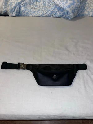 Svart magväska från BOSS Parfums - Snygg svart magväska från BOSS Parfums i slätt syntetmaterial. Väskan har ett metallspänne med logga, dragkedja upptill och ett coolt B-emblem framtill. Perfekt för att bära det viktigaste och ger en clean look till din outfit.