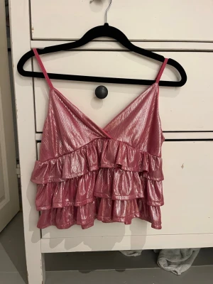 Glittrig rosa volangtop från SHEIN - Säljer en glittrig rosa topp från SHEIN i storlek XS. Toppen har tunna axelband och flera lager volanger som ger en riktigt festlig känsla. Materialet är skimrande och fångar ljuset snyggt. Perfekt för dig som vill sticka ut!