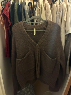 Brun stickad kofta från Kay/Day - Mysig brun stickad kofta från Kay/Day med två stora fickor framtill och v-ringad hals. Koftan har knappar och ribbade detaljer vid ärmslut och nederkant. Perfekt för lager på lager under kyliga dagar.