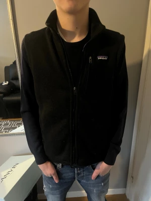 Svart fleeceväst från Patagonia - Säljer en svart fleeceväst från Patagonia med hög krage och dragkedja framtill. Västen har en bröstficka med dragkedja och diskret logga på vänster sida. Perfekt att slänga över en hoodie eller tröja för extra värme.
