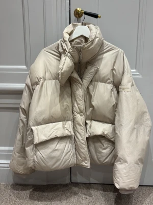 Beige dunjacka med hög krage - Säljer en beige dunjacka med oversized passform och hög krage. Jackan har stora fickor framtill och stängs med dragkedja och tryckknappar. Perfekt för kalla dagar och riktigt snygg till både jeans och sneakers.