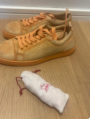 Beige mockasneakers från Louboutin - Snygga sneakers från Christian Louboutin i beige mocka med orange detaljer och snörning. Skorna har den ikoniska röda sulan och rund tå. Perfekta för dig som vill ha något unikt och exklusivt i garderoben.