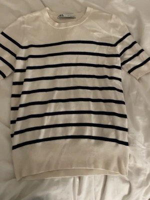 Randig vit t-shirt från zara - Säljer en vit t-shirt från zara med marinblå horisontella ränder. Klassisk rak passform och rund halsringning. Materialet känns mjukt och svalt, perfekt för varmare dagar. Enkel och stilren design som funkar till det mesta.