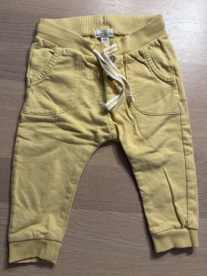 Gula mjukisbyxor med snörning - Söta gula mjukisbyxor från Newbie i storlek 68. Byxorna har resår i midjan, snörning framtill och muddar vid bensluten. Perfekta för chill dagar och lek. Mjukt bomullsmaterial som känns skönt mot huden.