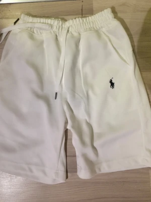 Vita shorts från Polo Ralph Lauren - Snygga vita shorts från Polo Ralph Lauren med elastisk midja och snörning. Klassisk svart logga broderad på benet. Tillverkade i mjukt material som känns skönt mot huden. Perfekta för sommardagar och chill häng.
