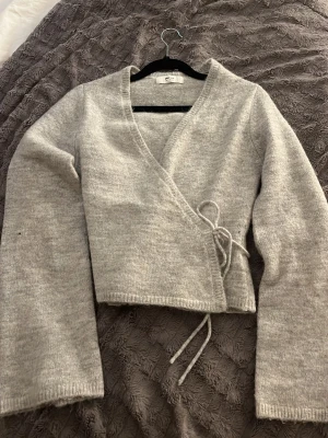 Grå omlottkofta med knyt - Mysig grå omlottkofta från Gina Tricot med knyt i sidan. Koftan är stickad, har lång ärm och croppad passform. Perfekt att slänga på sig över ett linne eller t-shirt för en chill look.