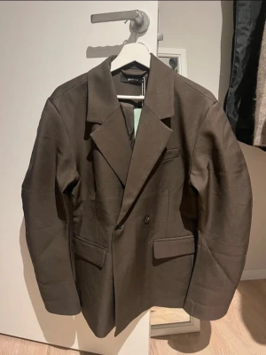 Brun kavaj från Gina Tricot - Snygg brun kavaj från Gina Tricot med klassisk dubbelknäppning och breda slag. Rak passform och långa ärmar. Perfekt att styla över en t-shirt eller skjorta för en clean look.