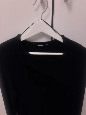 Svart ribbad topp från BikBok XL - Säljer en svart långärmad topp från BikBok i storlek XL. Toppen är ribbad och har en unik asymmetrisk detalj vid halsringningen som ger en cool twist. Materialet är mjukt och stretchigt. Helt o använde utan prislapp