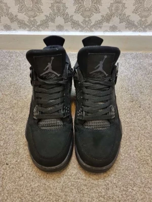 Svarta black cats/ Nike Air Jordan 4 sneakers - Säljer ett par helsvarta Nike Air Jordan 4 sneakers med klassisk siluett och coola meshdetaljer på sidorna. Skorna har snörning, platt sula och är tillverkade i mocka och syntet. Perfekta för dig som vill ha en stilren och ikonisk look.