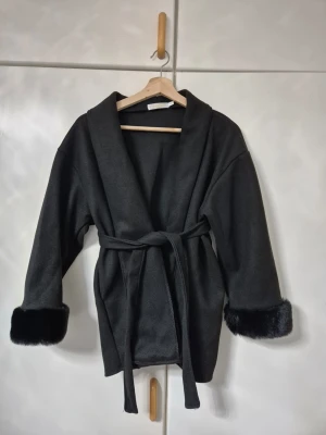 Svart kappa med fuskpälsdetaljer - Snygg svart kappa med bred krage och bälte i midjan. Ärmarna har lyxiga fuskpälsdetaljer vid muddarna som ger en extra touch. Perfekt för dig som vill ha en stilren och trendig look under kyliga dagar.
