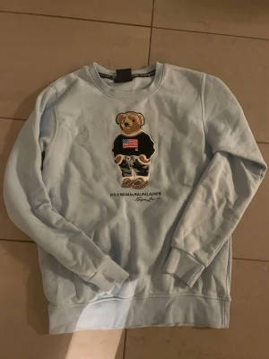 Ljusblå Polo Bear sweatshirt Ralph Lauren - Ljusblå sweatshirt från Polo Ralph Lauren med ikoniska Polo Bear broderad framtill. Tröjan har rund hals, långa ärmar och är gjord i mjuk bomull. Perfekt för dig som vill ha en chill och snygg look med en twist.