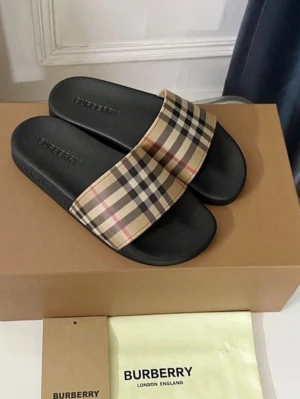 Burberry sandaler med rutigt mönster -  riktiga Burberry tofflor med klassiskt beige, svart och rött rutigt mönster över foten. Sulan är svart och har Burberry-loggan präglad. Perfekta för sommaren och ger en lyxig touch till din outfit.