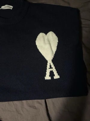 Mörkblå stickad tröja från Ami Paris - Säljer en mörkblå stickad tröja från Ami Paris med deras ikoniska vita hjärta och A på bröstet. Tröjan har rund halsringning, ribbade muddar och lång ärm. Perfekt för dig som vill ha en clean men ändå statement look.