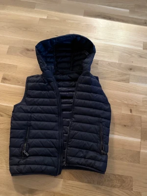 Massimo dutti väst  - Snygg mörkblå dunväst med quiltad design och praktisk huva. Västen har dragkedja framtill och två fickor med dragkedja. Perfekt att slänga på över en hoodie för extra värme under kyliga dagar. Defekt en dragkedjan är trasig och ett jätte litet hål på höger sida kan skicka bild vid önskan