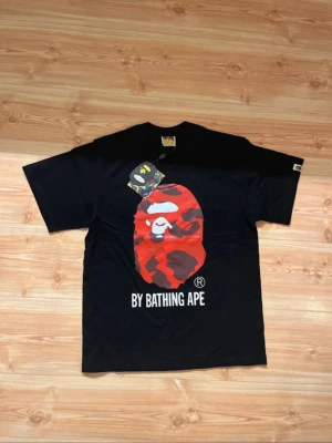 Svart BAPE t-shirt med röd camo print - Svart t-shirt från BAPE med stort rött camo-apansikte och texten 'BY BATHING APE' på bröstet. Klassisk rund hals, korta ärmar och liten BAPE-logga på ärmen. Tillverkad i mjuk bomull för skön passform.
