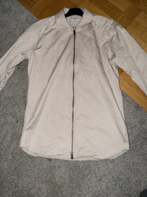 Beige overshirt från John Henric - Säljer en stilren beige overshirt från John Henric. Jackan har rak passform, klassisk krage och hel dragkedja framtill. Tillverkad i mjuk bomull och passar perfekt för lager på lager. Enkel och clean look som funkar till många outfits.