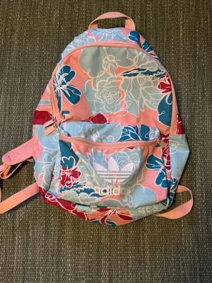Blommig ryggsäck från Adidas - Säljer en färgglad ryggsäck från Adidas med blommigt mönster i rosa, ljusblått, rött och vitt. Väskan har ett stort huvudfack, en mindre ficka framtill med dragkedja och justerbara axelband. Perfekt för dig som vill sticka ut med en unik och snygg väska.