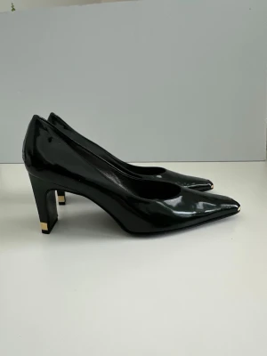 Svarta lackpumps med gulddetaljer - Snygga svarta pumps i äkta skinn med spetsig tå och fyrkantig klack. Gulddetaljer på både tå och klack ger en lyxig känsla. Perfekta för dig som vill sticka ut med en klassisk men ändå unik look.