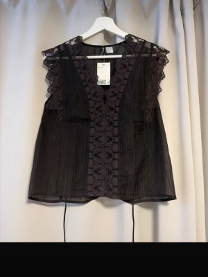 Ny populär Svart blus med spets från H&M - Supersöt svart blus från Divided i transparent tyg med broderade spetsdetaljer längs v-ringningen och axlarna. Blusen har knytband i midjan och är ärmlös, perfekt för att lyfta vilken outfit som helst. HELT NY och helt slutsåld på hemsidan !!
