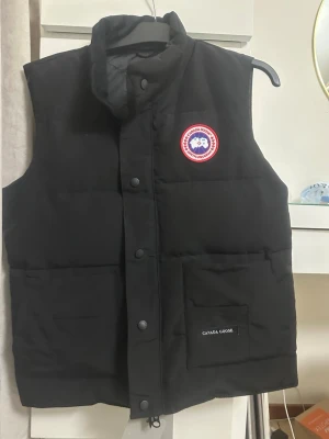 Canada Goose  - Säljer en svart dunväst från Canada Goose med hög krage och tryckknappar framtill. Västen har två stora fickor och en tydlig Canada Goose-logga på bröstet. Materialet är slitstarkt och västen är perfekt för kyliga dagar. Klassisk och stilren design. Oanvänt 