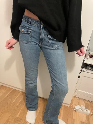 Blå bootcut jeans med fickdetaljer - Säljer ett par ljusblå jeans med bootcut passform och snygga fickor med lock och knapp framtill. Jeansen har normal midja och klassisk femficksdesign. Materialet är mjukt denim som sitter skönt och följer kroppen.har en liten slitning vid fickan💕