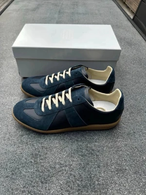 Mörkblå sneakers med mockadetaljer - Säljer ett par stilrena mörkblå sneakers med ovandel i mocka och läder, beige innersula och vita snören. Skorna har en klassisk låg siluett och gummisula som ger en retrovibe. Perfekta för dig som gillar minimalistisk och clean stil. 