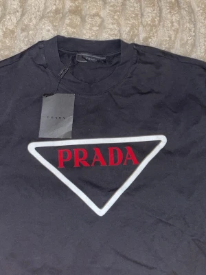 Svart t-shirt från Prada med logga - Svart t-shirt från Prada med ikonisk triangel-logga framtill i vitt och rött. Klassisk rund halsringning och korta ärmar. Tillverkad i 100% bomull för en skön och avslappnad känsla. Perfekt för dig som vill ha en clean och stilren look.