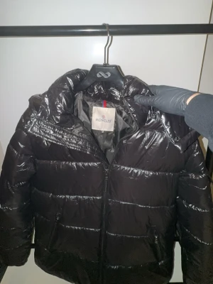 Svart Moncler pufferjacka  - Svart pufferjacka från Moncler med glansig finish och klassisk logga på ärmen. Jackan har huva, dragkedja framtill och praktiska fickor. Insidan har en cool serietidningsetikett. Perfekt för dig som vill ha en varm och snygg jacka med streetkänsla.