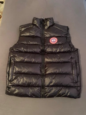 Canada Goose väst - Säljer en svart dunväst från Canada Goose med klassisk logga på bröstet. Västen har hög krage, dragkedja framtill och två stora fickor. Den quiltade designen ger en snygg och sportig look, perfekt för kyliga dagar.