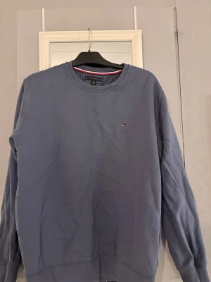 Blå sweatshirt från Tommy Hilfiger XL - Säljer en blå sweatshirt från Tommy Hilfiger i storlek XL. Tröjan har rund halsringning med ribbade muddar och en liten broderad logga på bröstet. Perfekt för dig som gillar klassisk och stilren streetwear.