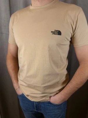 Beige t-shirt från The North Face - Snygg beige t-shirt från The North Face med klassisk logga tryckt i svart på bröstet. T-shirten har rund halsringning och korta ärmar. Tillverkad i mjuk bomull som känns skön mot huden. Perfekt till jeans eller shorts för en clean och enkel look.