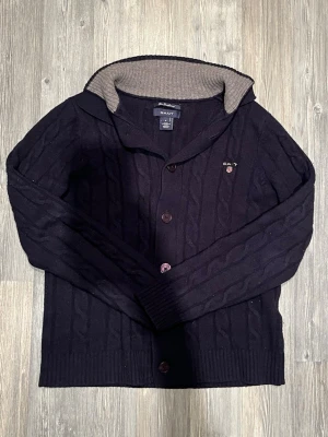 Mörkblå kabelstickad kofta från GANT - Säljer en mörkblå kabelstickad kofta från GANT i mjuk lammull. Koftan har knappar framtill, ribbade muddar och en grå ribbstickad insida på kragen. Broderad GANT-logga med sköld på bröstet. Perfekt för kyliga dagar!