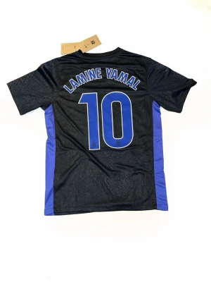 Svarta Nike fotbollströja FC Barcelona - Säljer en svart och blå FC Barcelona fotbollströja med Lamine Yamal och nummer 10 på ryggen. Tröjan har lila detaljer, FCB-logga och Spotify-logga på bröstet. Materialet är lätt polyester med subtilt mönster och korta ärmar. Perfekt för dig som älskar Barca och vill ha en snygg matchtröja.