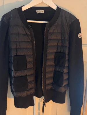 Svart kofta från Moncler - Säljer en svart quiltad kofta från Moncler med ribbade muddar och dragkedja framtill. Koftan har puffade partier på framsidan, fyra fickor och Moncler-logga på ärmen. Perfekt för lager på lager under höst och vinter.