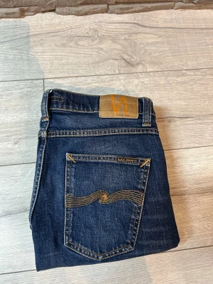 Mörkblå Nudie Jeans, W31 L32 - Säljer ett par mörkblå jeans från Nudie Jeans med klassisk femficksmodell och snygga kontrastsömmar. Jeansen har en rak passform och ikonisk broderad bakficka. Tillverkade i slitstarkt denim med en cool tvättad look.