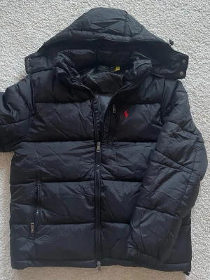 Svart pufferjacka från Polo Ralph Lauren - Säljer en svart pufferjacka från Polo Ralph Lauren med huva och dragkedja. Jackan har det klassiska röda Polo-logot broderat på bröstet och två fickor med dragkedja. Perfekt för kalla dagar och riktigt snygg till streetwear-stilen.