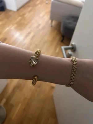 Guldig armband med stor kristall - Säljer ett riktigt snyggt armband i guldton med en stor, droppformad kristall i mitten. Armbandet har en cool struktur med små guldfärgade kulor längs hela bandet och är öppet baktill för enkel påtagning. Perfekt för dig som vill sticka ut lite extra.