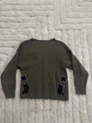 Olivgrön stickad tröja med snörning - Säljer en olivgrön stickad tröja med långa ärmar och coola snörningsdetaljer i mörkblått på sidorna. Tröjan har en klassisk rund halsringning och ribbade muddar vid ärmslut och nederkant. Perfekt för lager på lager under hösten.