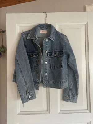 Blå jeansjacka från Nelly - Säljer en klassisk blå jeansjacka från Nelly i storlek XS. Jackan har två bröstfickor med knappar, silverfärgade knappar framtill och vid ärmslut samt en rak, något croppad passform. Perfekt att slänga över en hoodie eller t-shirt för en chill look.