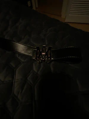 Svart amiri  bälte med stor metallspänne - Säljer ett svart bälte  med ett stort, coolt metallspänne i form av ett M. Bältet har vit söm längs kanterna och passar perfekt till jeans kvaliten är jätte bra storlek 110cm+
