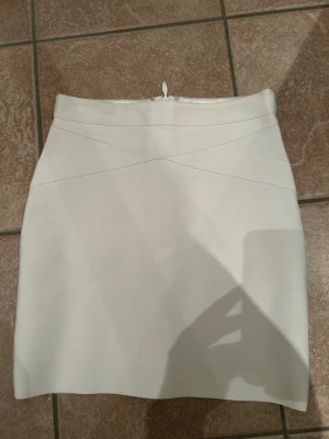 Vit kortkjol från BikBok - Säljer en stilren vit kortkjol från BikBok i storlek S. Kjolen har en figurnära passform, diskreta sömdetaljer framtill och dragkedja bak. Materialet känns mjukt och har en lätt glansig finish på insidan.