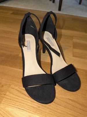 Svarta klackar - Stilrena svarta sandaler från Nelly med öppen tå och smal rem över foten. Skorna har en elegant smal klack och justerbart spänne vid ankeln. Perfekta för dig som vill ha en enkel men snygg sko till många olika outfits.