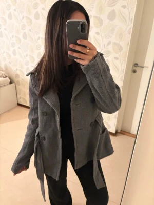 Grå kappa från H&M med bälte - Säljer en grå kappa i mjuk manchester med bred krage, dubbelknäppning och knytskärp i midjan. Jackan har stora fickor framtill och långa ärmar. Perfekt för dig som gillar en klassisk men chill stil. Kappan har dock slitage i form av hål vid armhålorna, men detta är inget som syns från utsidan och märks inte när jackan bärs. Den är i storlek 36 men passar för mig som bär XXS/XS, alltså även 32/34.