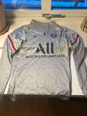 Grå PSG x Jordan träningströja XS - Säljer en grå långärmad PSG träningströja från Jordan med mörkblå prickigt mönster och röda/blå detaljer på ärmarna. Tröjan har half zip, Paris Saint-Germain-logga på bröstet och är i slim fit. Perfekt för dig som gillar fotboll och streetwear.