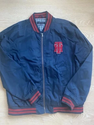 Blå bomberjacka Tommy Hilfiger - Snygg mörkblå bomberjacka från Tommy Hilfiger med röda och svarta ränder på krage, ärmslut och midja. Jackan har dragkedja framtill, broderad TH-logga på bröstet och rutigt innerfoder. Perfekt för dig som vill ha en klassisk college-look. I storlek L