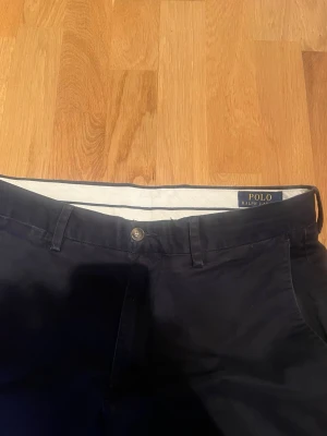 Mörkblå chinos från Polo Ralph Lauren - Snygga mörkblå chinos från Polo Ralph Lauren med stretch och slim fit. Klassisk design med bälteshällor, knappgylf och två fickor fram samt två bakfickor med knapp. Perfekta för en clean och stilren look. Materialet är mjukt och bekvämt, troligtvis bomull med lite stretch.