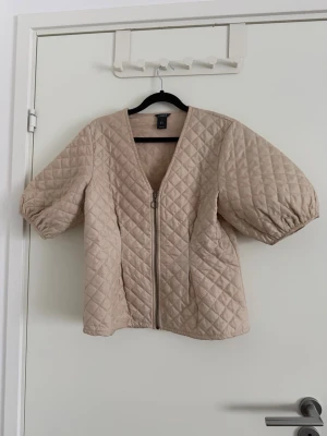 Beige quiltad jacka från Lindex - Quiltad beige jacka från Lindex med korta puffärmar och dragkedja framtill. Jackan har ett rutigt mönster och en loose passform som ger en chill vibe. Perfekt att slänga på sig över en hoodie eller stickad tröja. Enkel och stilren design som funkar till många outfits. Använd fåtal gånger.