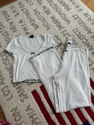 Vit randig pyjamas set från Gina Tricot - Supersöt vit pyjamas set från Gina Tricot i ribbad bomull med tunna ränder och små hålmönster. Svarta volangdetaljer och en liten rosett framtill ger en gullig vibe. Perfekt att styla lager på lager eller till jeans för en chill look.